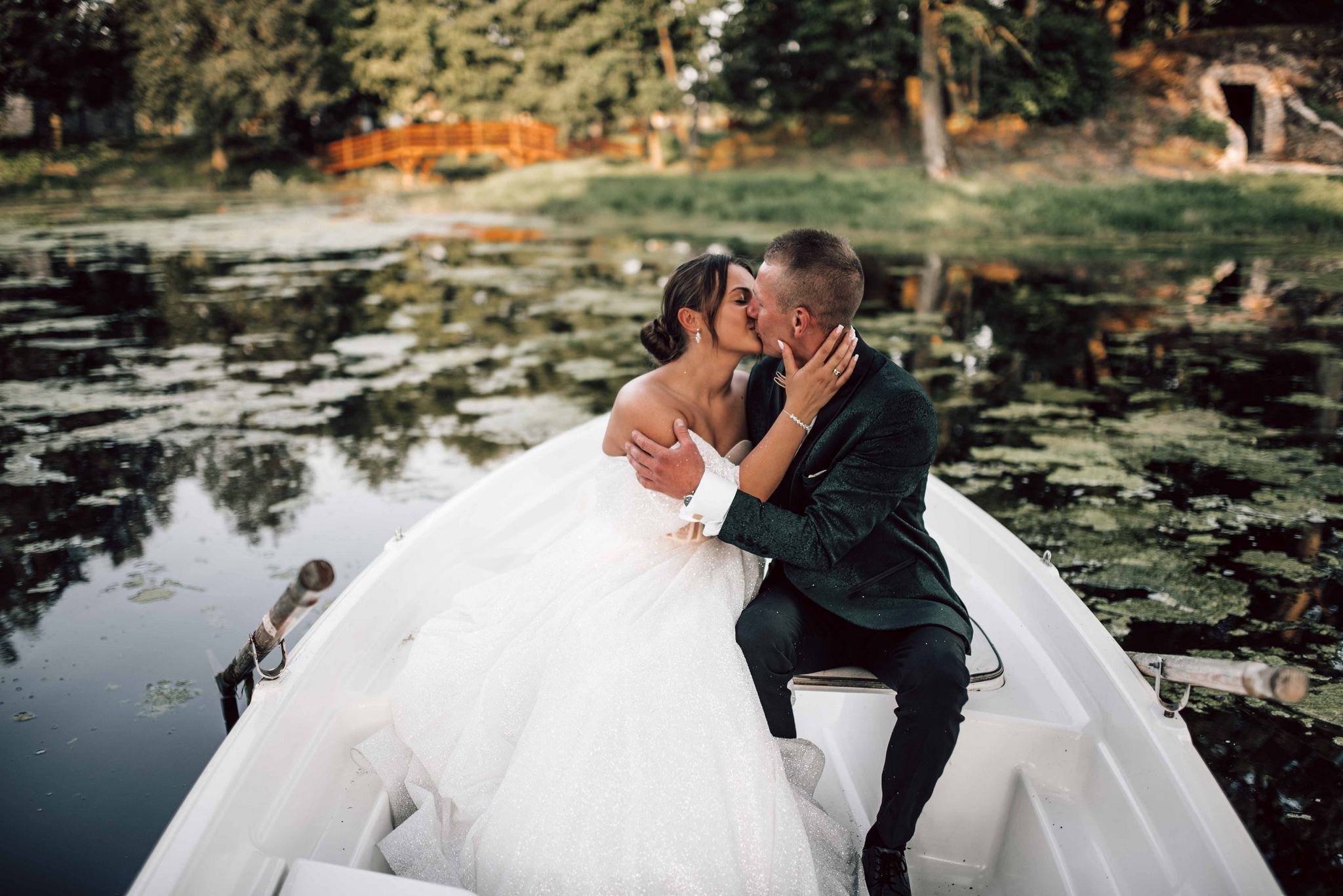 Pocałunek nowożeńców w łódce na jeziorze, ona w błyszczącej sukni, on w ciemnozielonym garniturze. W tle zalesiony brzeg i drewniany mostek.