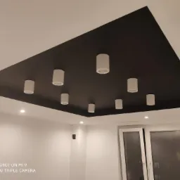 Część sufitu z czarną, kwadratową wnęką, w której zamontowano osiem białych, cylindrycznych lamp punktowych, na tle białych ścian i okna balkonowego.