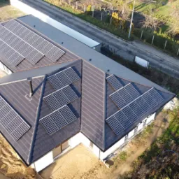 Widok z g&oacute;ry na dom z ciemną dach&oacute;wką, na kt&oacute;rej zamontowane są panele słoneczne, obok domu widoczny plac budowy i droga.