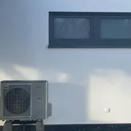 Zewnętrzna jednostka klimatyzacyjna, prawdopodobnie typu split, ustawiona na betonowych podstawach przy białej ścianie budynku z małym, prostokątnym oknem w górnej części kadru. Widoczne delikatne...