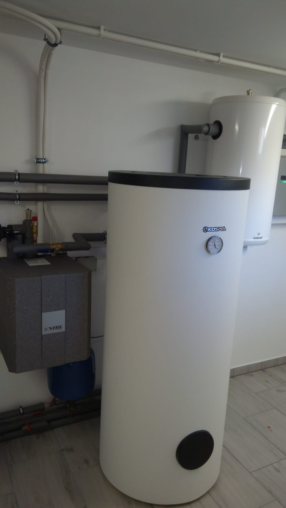 Biały zbiornik buforowy Kospel z manometrem i czarną zaślepką u dołu, obok szara jednostka Nibe z niebieskim elementem, rury instalacyjne na ścianie, jasne pomieszczenie z płytkami na podłodze.
