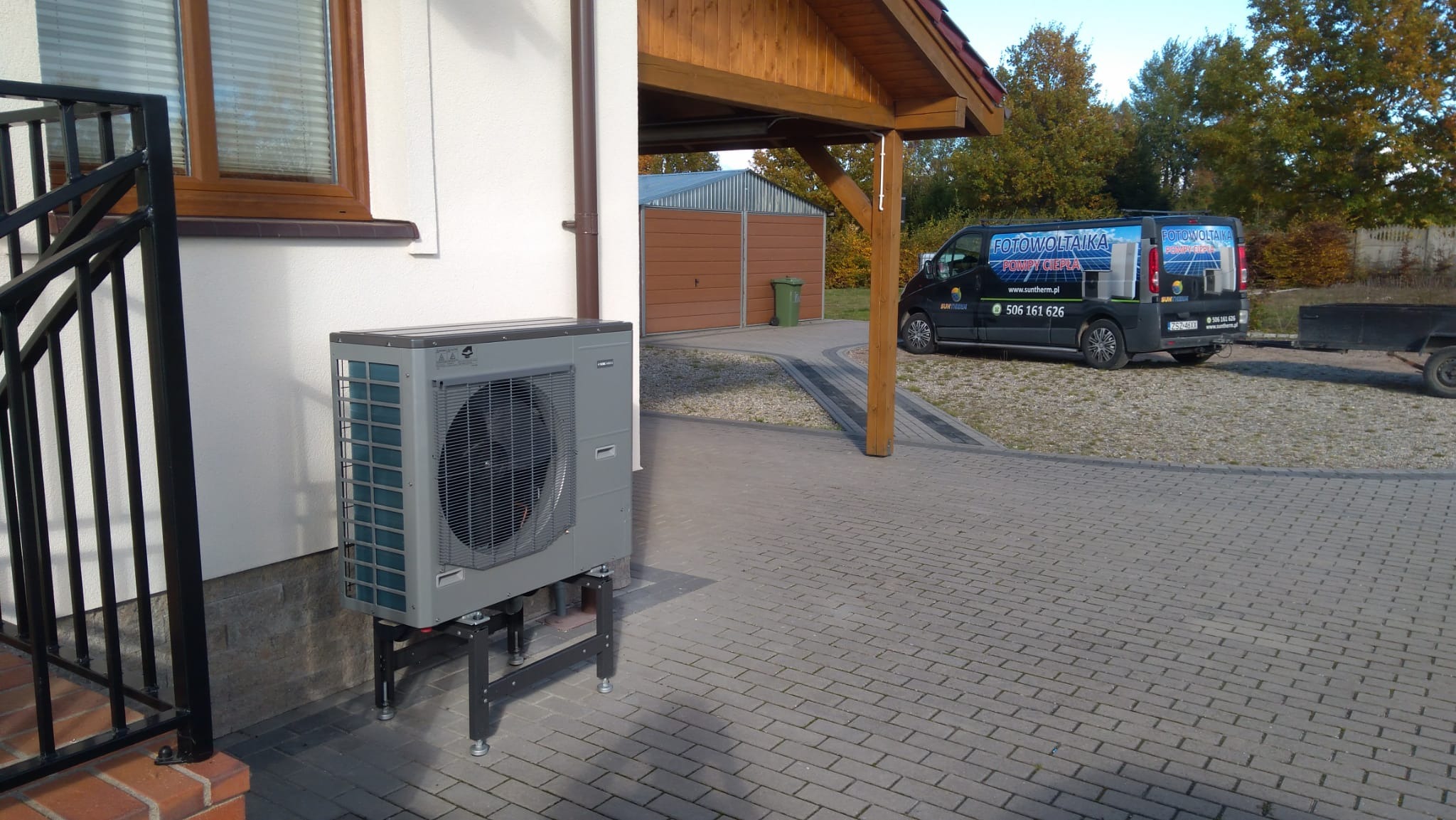 Szczecinek: Szara jednostka zewnętrzna urządzenia grzewczego na metalowym stelażu, zamontowana przy ścianie budynku z brązowymi oknami, na tle podjazdu z kostki brukowej i zaparkowanego busa...