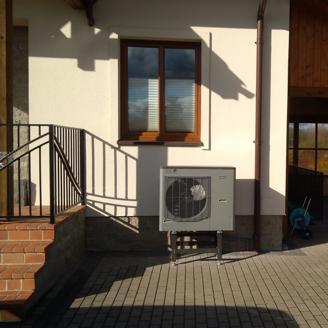 Szara zewnętrzna jednostka pompy ciepła zamontowana na czarnym metalowym stojaku, ustawiona przy białej ścianie budynku z brązowym oknem i rynną, widoczne ceglane schody z czarną barierką i kostka...
