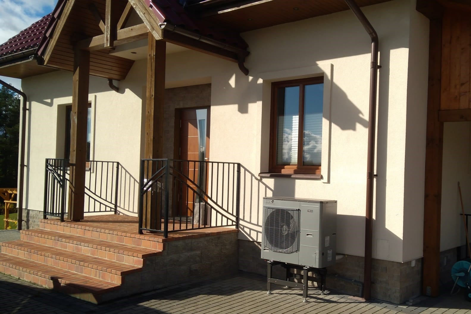 Szara jednostka zewnętrzna urządzenia grzewczego na metalowym stelażu, ustawiona obok jasnego budynku z brązową stolarką okienną i schodami wejściowymi z czarną balustradą.