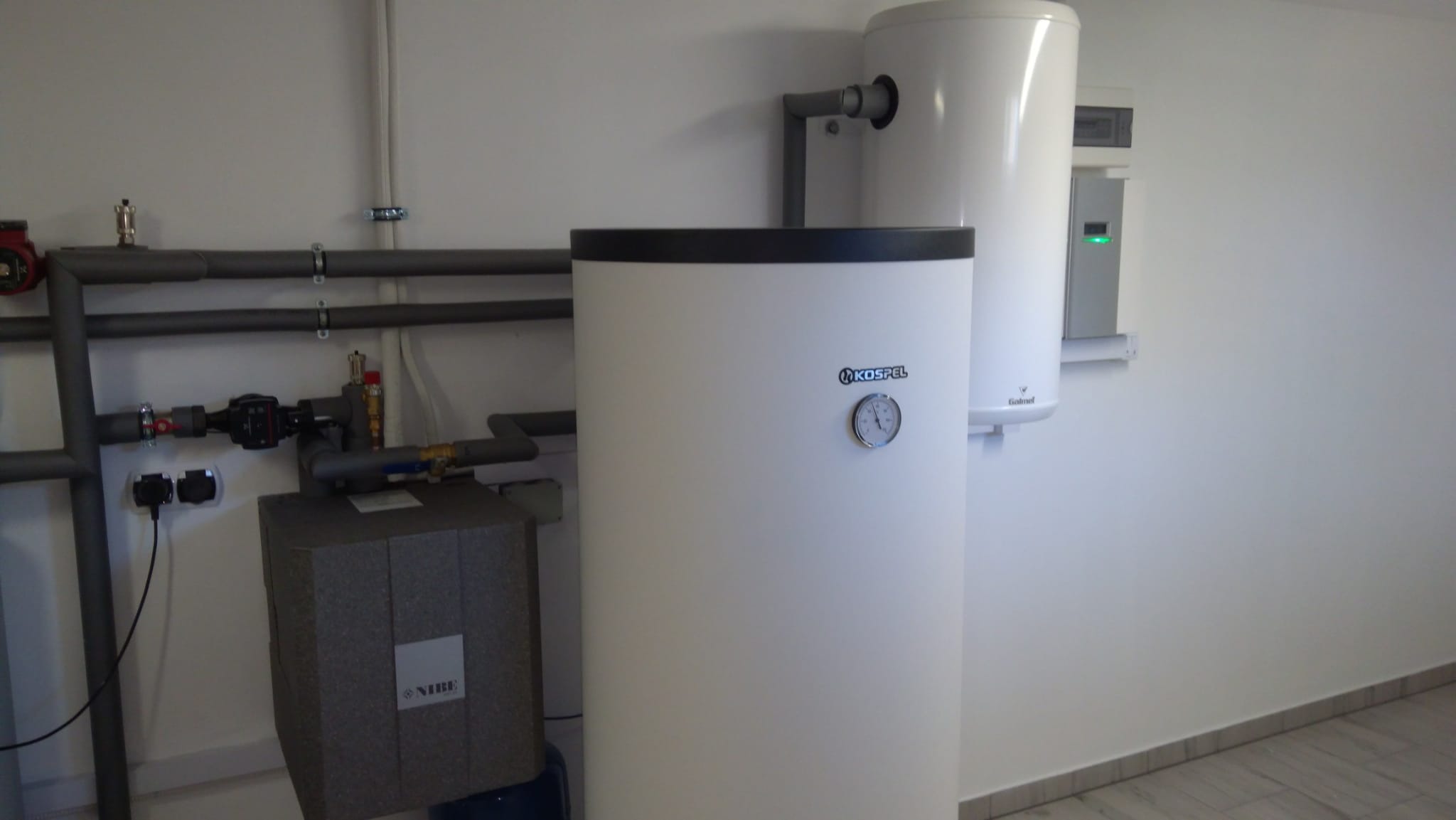 Biały zbiornik Kospel z czarnym obramowaniem u góry, obok mniejszy zbiornik Galmet z zieloną diodą, szare rury instalacyjne i blok NIBE na białej ścianie z szarą podłogą.