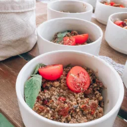 Rząd białych misek z kaszą gryczaną, suszonymi pomidorami, świeżymi pomidorami i listkiem bazylii, ustawionych na stole z jutowym bieżnikiem, obok koszyka z pieczywem.