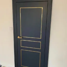 Eleganckie, ciemnogranatowe drzwi wewnętrzne z złotymi zdobieniami i klamką, widoczne na tle jasnej ściany i drewnianej podłogi