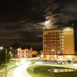 Nocny widok na wielopiętrowy hotel z logiem 'Gold Hotel' na szczycie, oświetlony księżycem przebijającym się przez chmury, z długimi smugami świateł samochodowych na pierwszym planie.