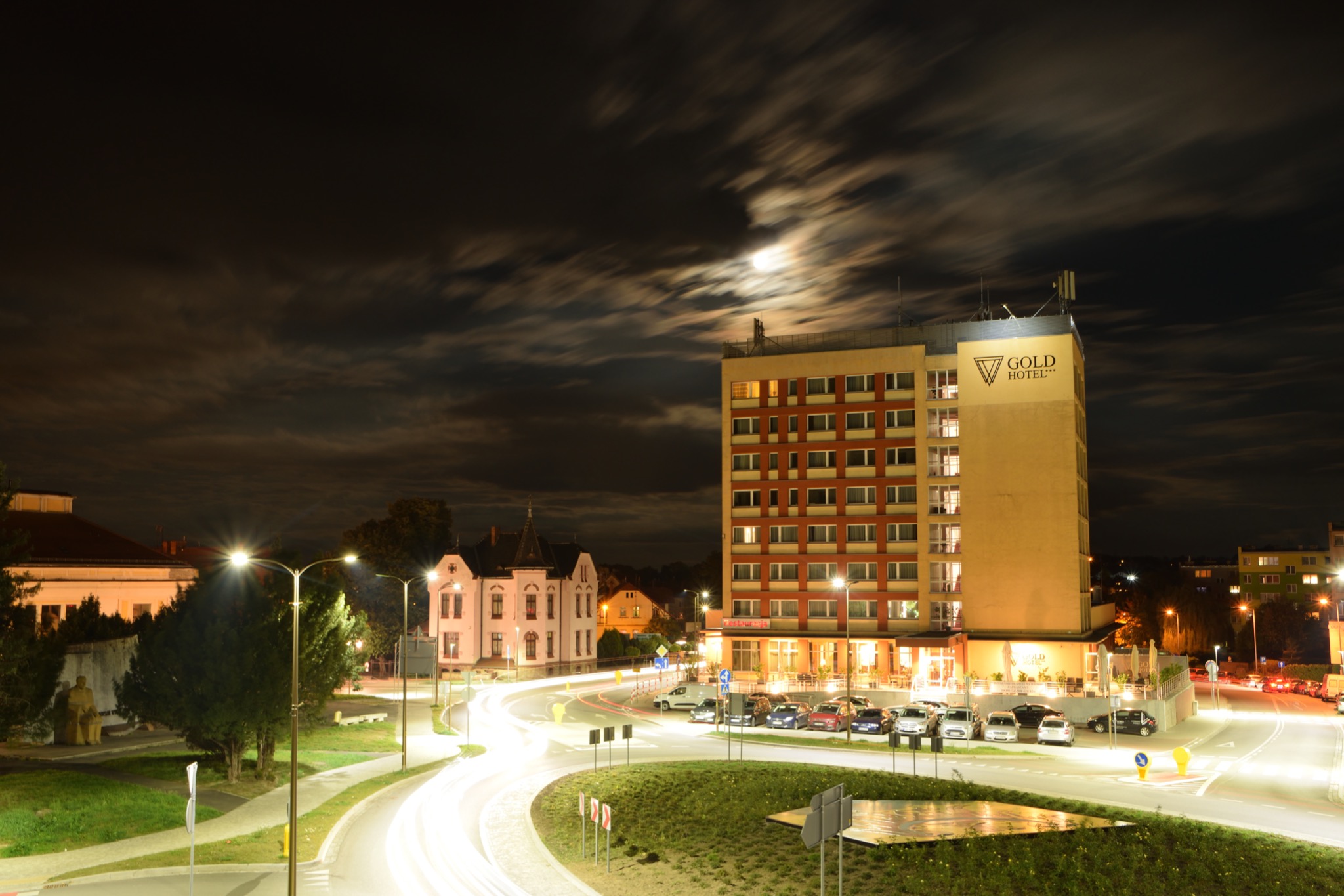 Nocny widok na wielopiętrowy hotel z logiem 'Gold Hotel' na szczycie, oświetlony księżycem przebijającym się przez chmury, z długimi smugami świateł samochodowych na pierwszym planie.