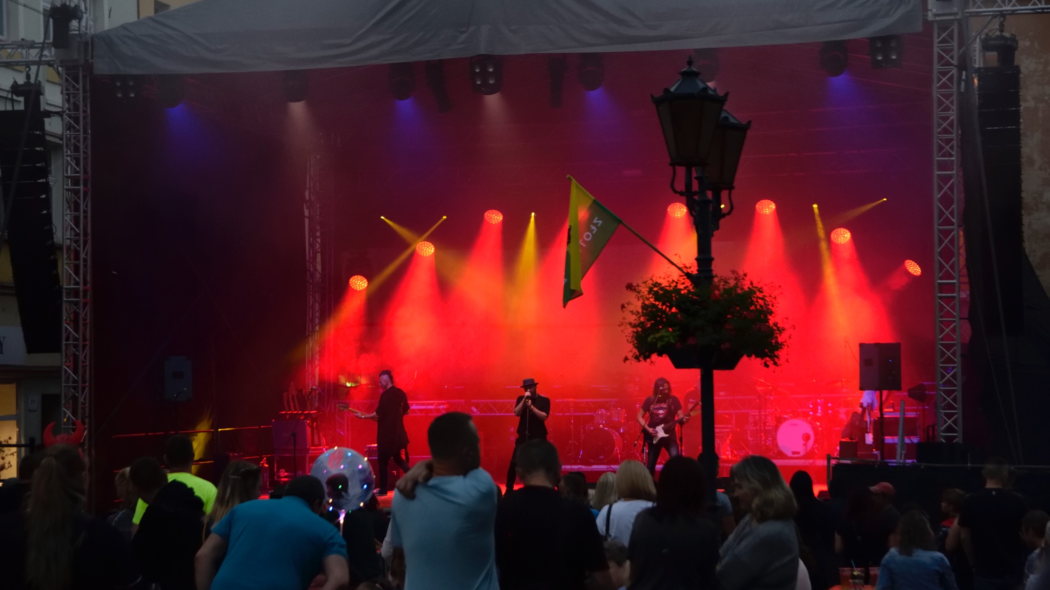 Zespół muzyczny na scenie oświetlonej czerwonymi i żółtymi reflektorami, widok zza publiczności, flaga i latarnia z kwiatami na pierwszym planie.