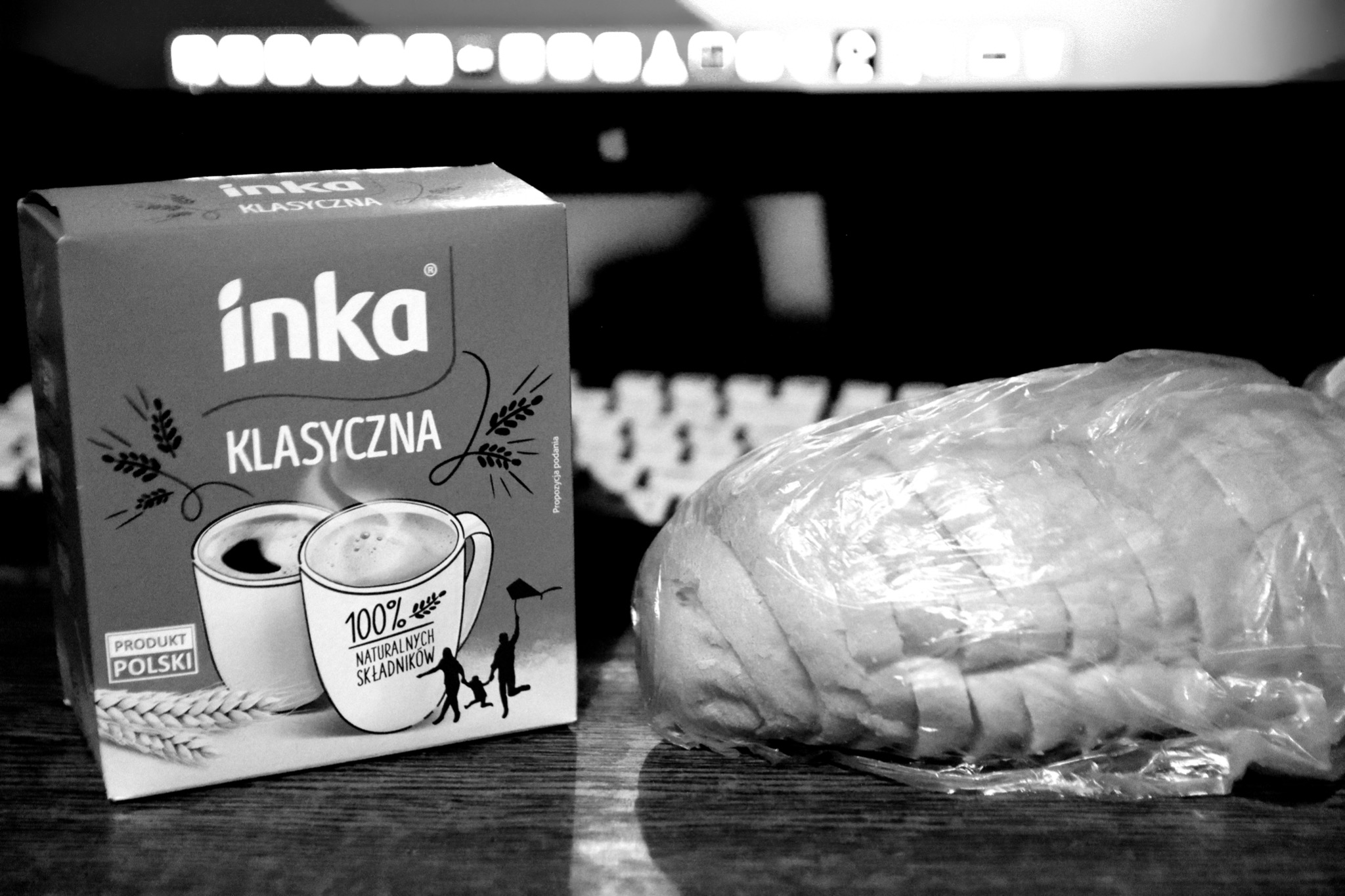 Pudełko kawy Inka Klasyczna obok zapakowanego chleba na tle klawiatury komputera i monitora.