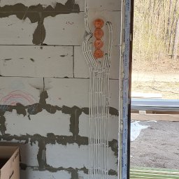 PRO-Instal - Instalacja elektryczna w trakcie montażu: białe przewody prowadzone w peszlach, wychodzące z pomarańczowych puszek podtynkowych, ułożone pionowo na ścianie z betonu komórkowego obok okna.