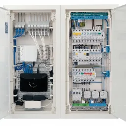 Otwarta szafa rozdzielcza z widocznymi bezpiecznikami, przewodami i urządzeniami sieciowymi, w tym routerem i zasilaczami, zamontowanymi wewnątrz białej obudowy.