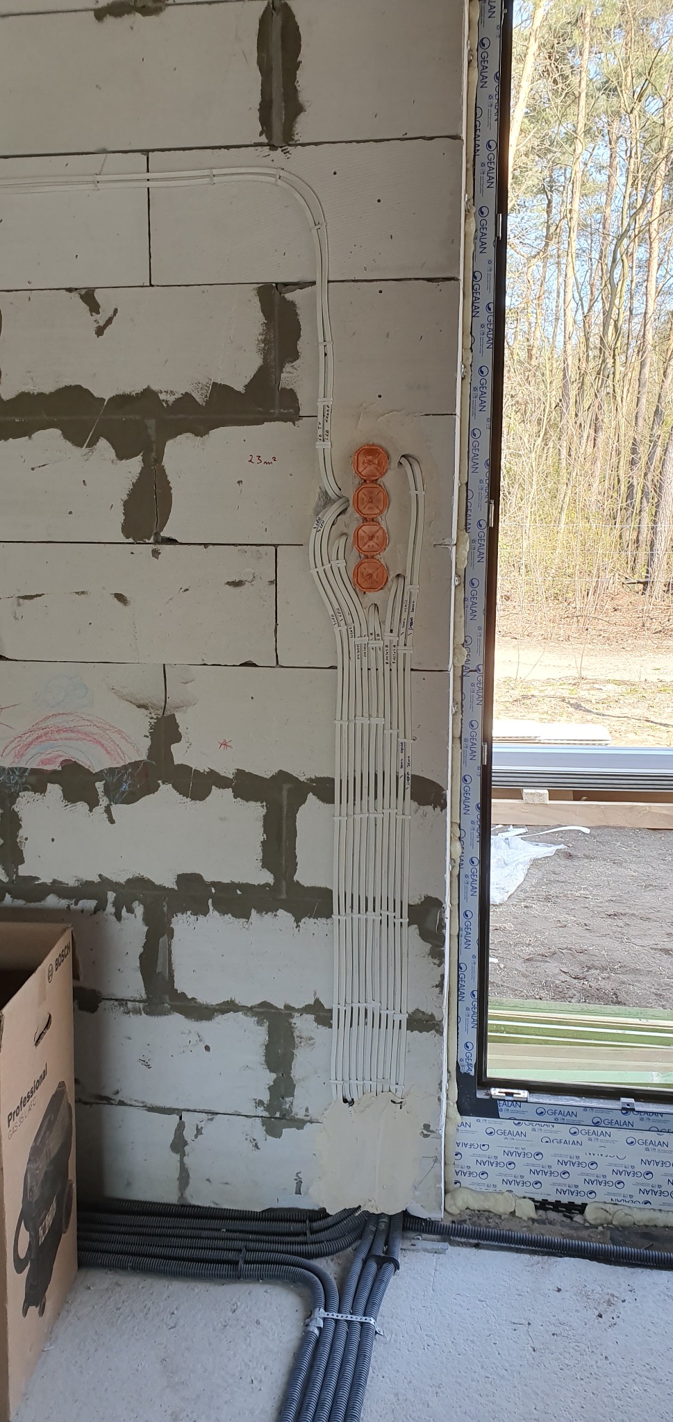 Instalacja elektryczna w trakcie montażu: białe przewody prowadzone w peszlach, wychodzące z pomarańczowych puszek podtynkowych, ułożone pionowo na ścianie z betonu komórkowego obok okna.