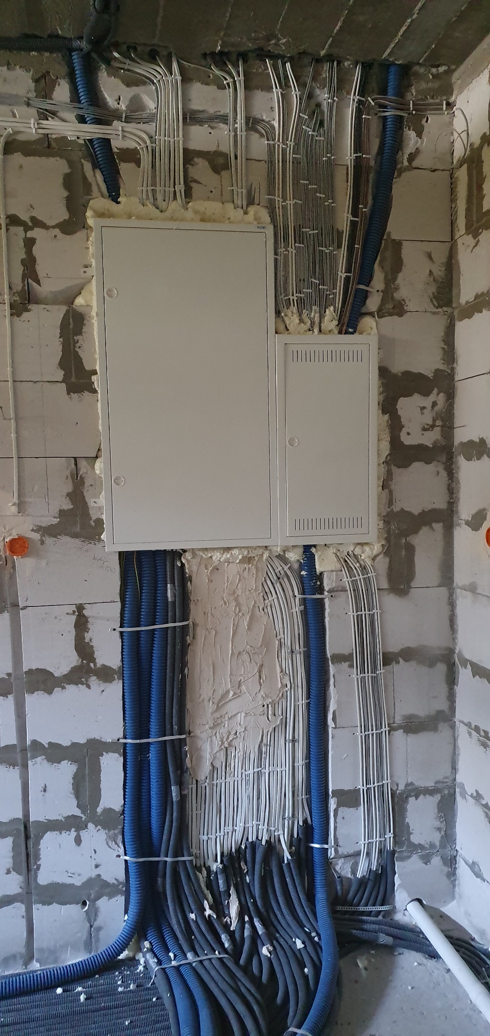 Rozdzielnia elektryczna natynkowa z licznymi przewodami w peszlach i kablami, montowana na ścianie z betonu komórkowego, zabezpieczona pianką montażową i gipsem.