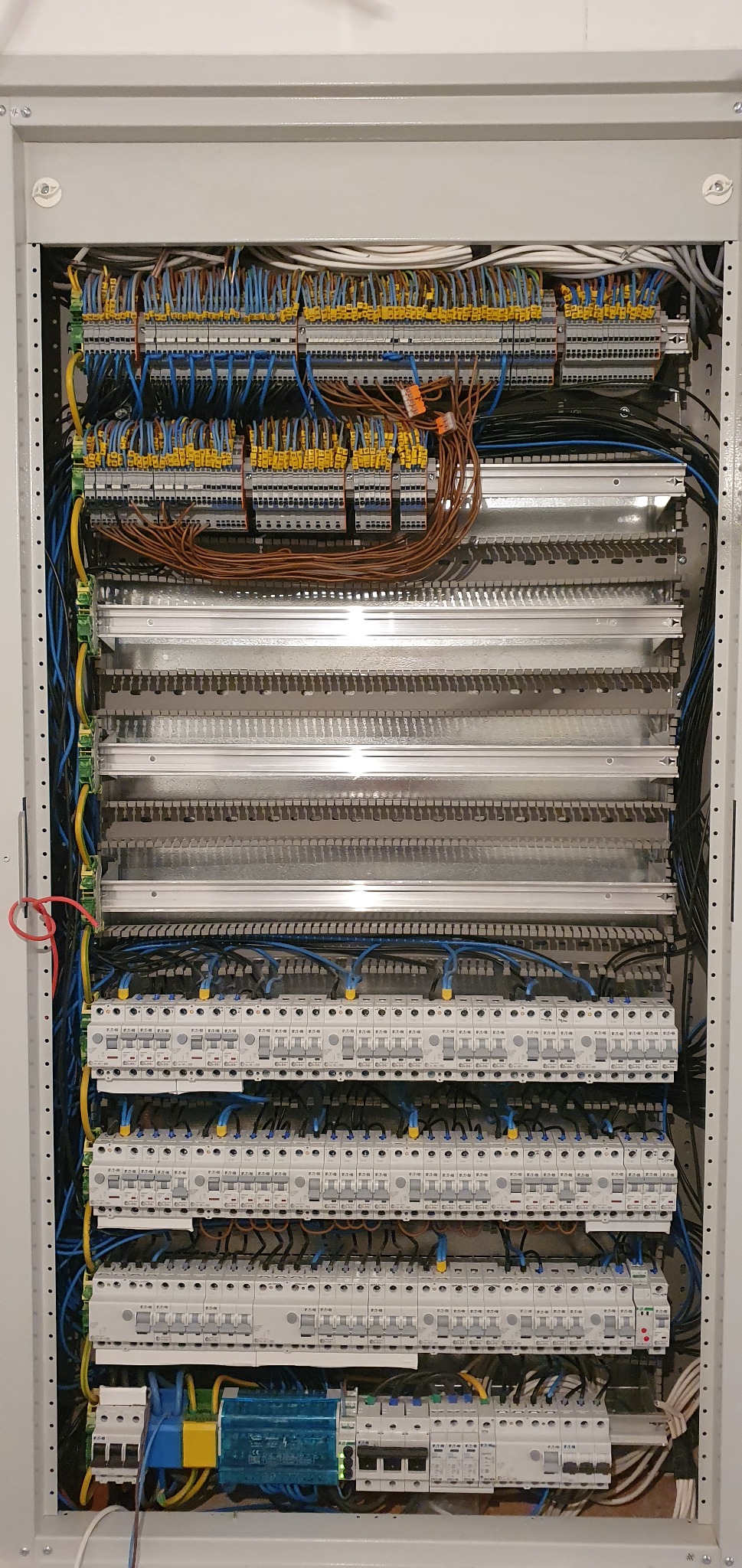 Rozdzielnia elektryczna w szarej obudowie, widoczne liczne bezpieczniki, przewody w różnych kolorach (niebieski, żółty, brązowy) oraz listwy zaciskowe z żółtymi oznaczeniami.