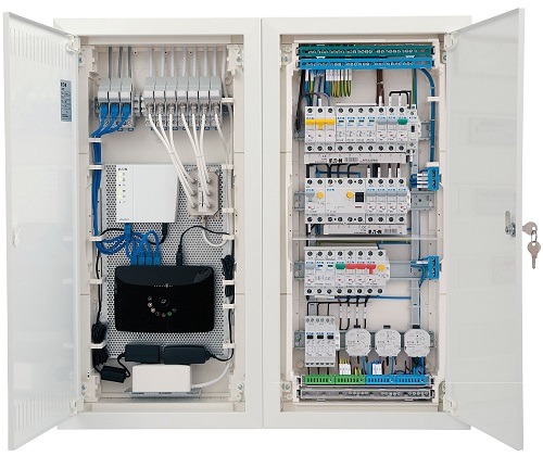 Otwarta szafa rozdzielcza z widocznymi bezpiecznikami, przewodami i urządzeniami sieciowymi, w tym routerem i zasilaczami, zamontowanymi wewnątrz białej obudowy.