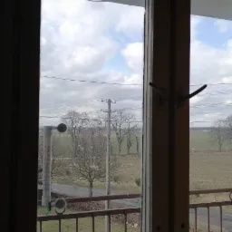 Widok przez brudne okno na zewnątrz, na balkon z metalową barierką, za którym rozciąga się wiejski krajobraz z polami, drogą i liniami energetycznymi, z termometrem przyklejonym do szyby.