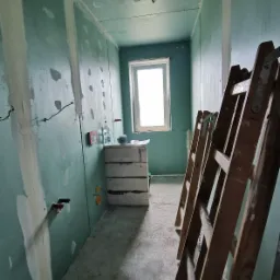 Wnętrze w trakcie prac przygotowawczych do wykończenia: ściany pokryte płytami gipsowo-kartonowymi z widocznymi łączeniami, niezamaskowane przewody elektryczne, drabina oparta o ścianę...