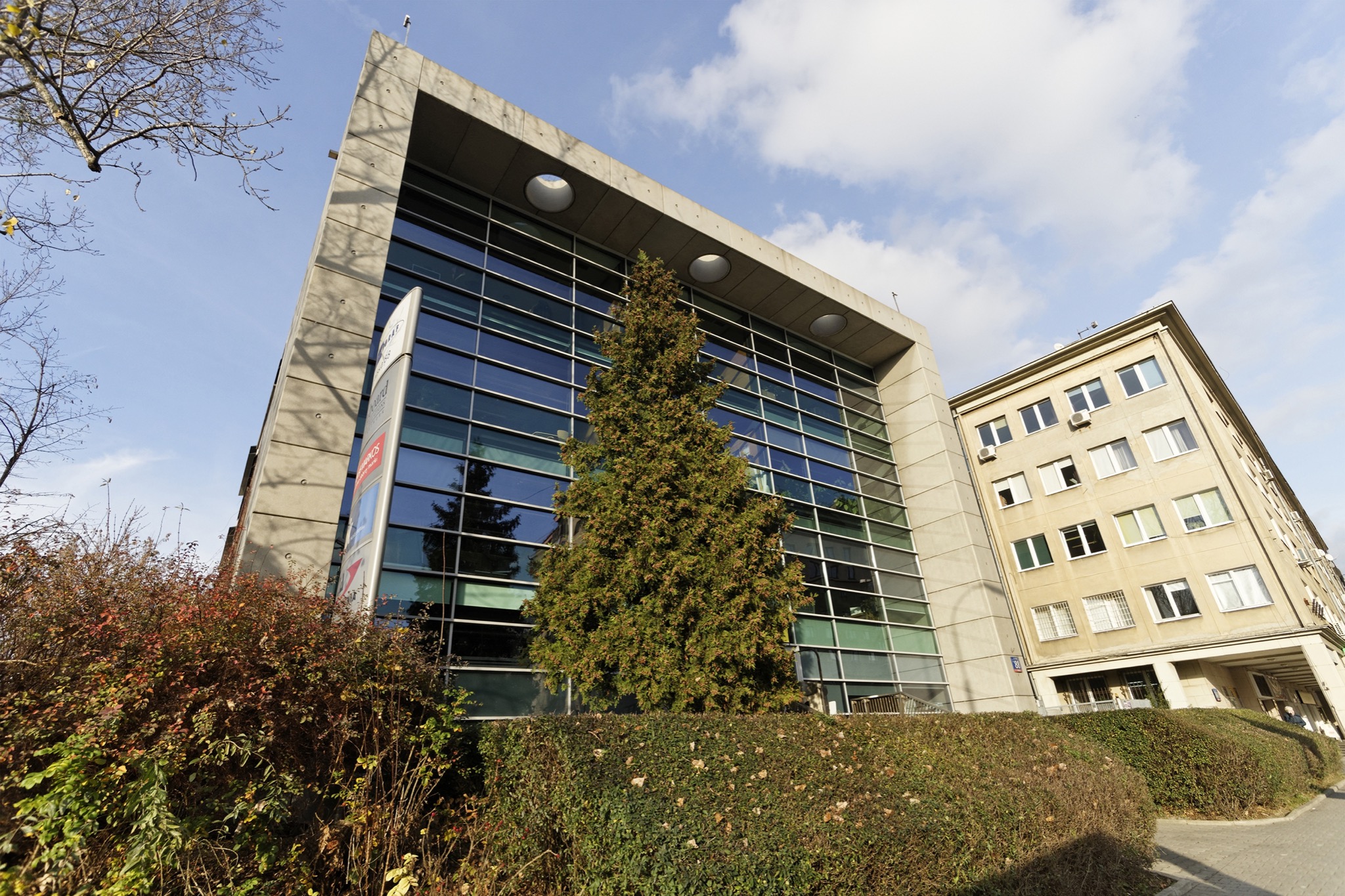 Nowoczesny budynek biurowy z przeszkloną fasadą i drzewem iglastym na pierwszym planie, widok z dołu na tle błękitnego nieba z chmurami.