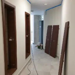 Wnętrze korytarza w trakcie montażu drzwi wewnętrznych z widocznymi ościeżnicami w ścianie i skrzydłami drzwiowymi opartymi o ścianę, zabezpieczone taśmą malarską ściany.