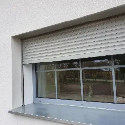 Szara roleta zewnętrzna opuszczona na oknie z szarymi ramami, widok z zewnątrz na białą elewację budynku.