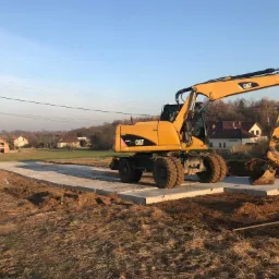 Żółta koparka kołowa CAT na placu budowy, stojąca na betonowych płytach, w tle domy jednorodzinne i błękitne niebo.