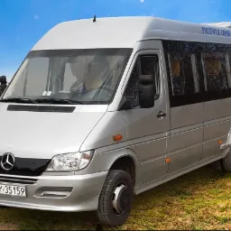 Srebrny bus Mercedes-Benz z napisem 'PRZEWÓZ OSÓB' i numerem telefonu na dachu, zaparkowany na trawiastym terenie z widokiem na góry w tle.