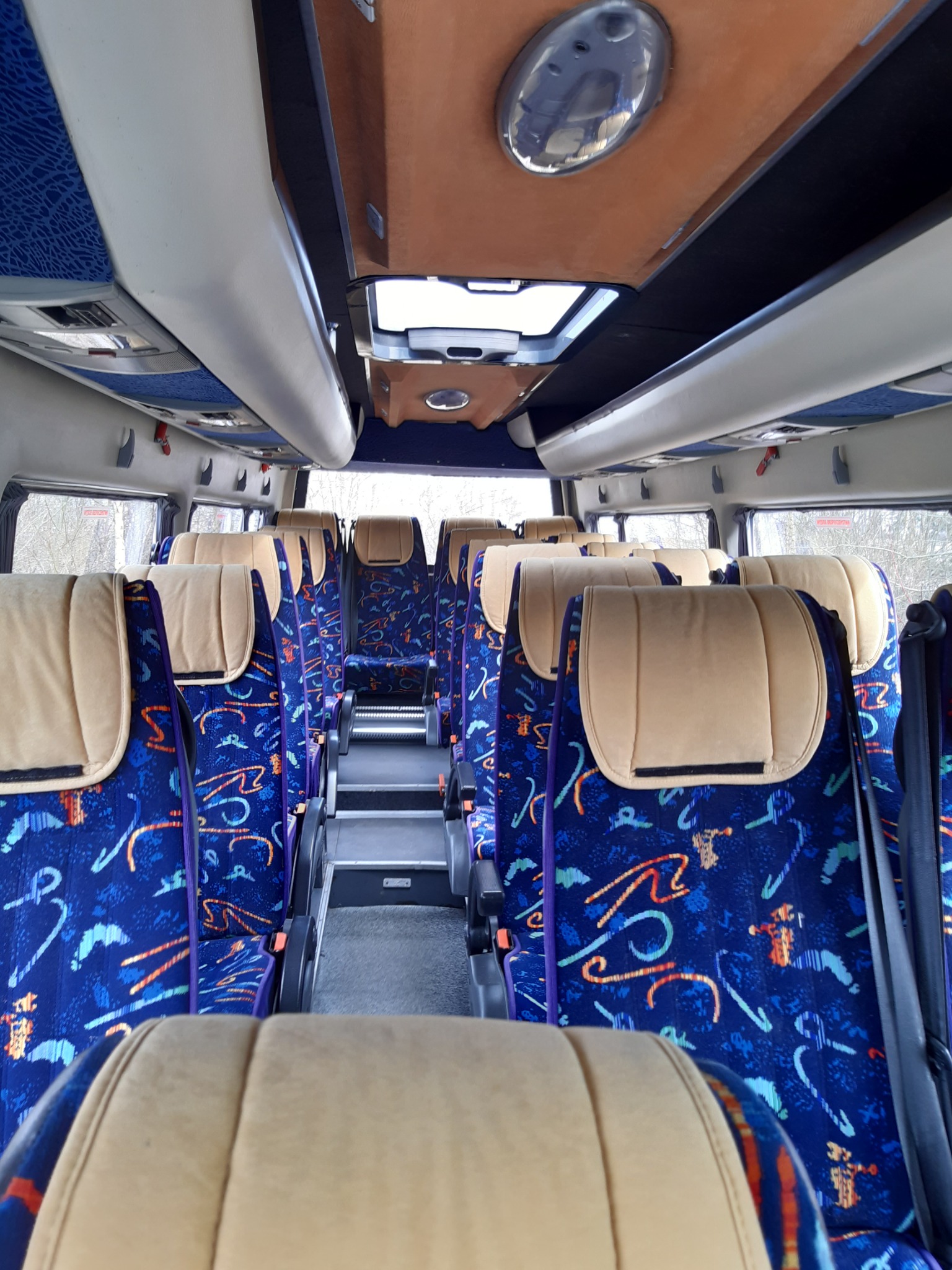 Wnętrze autobusu z niebieskimi siedzeniami w kolorowe wzory i beżowymi zagłówkami, widok w kierunku tyłu pojazdu, z oknem dachowym na górze.
