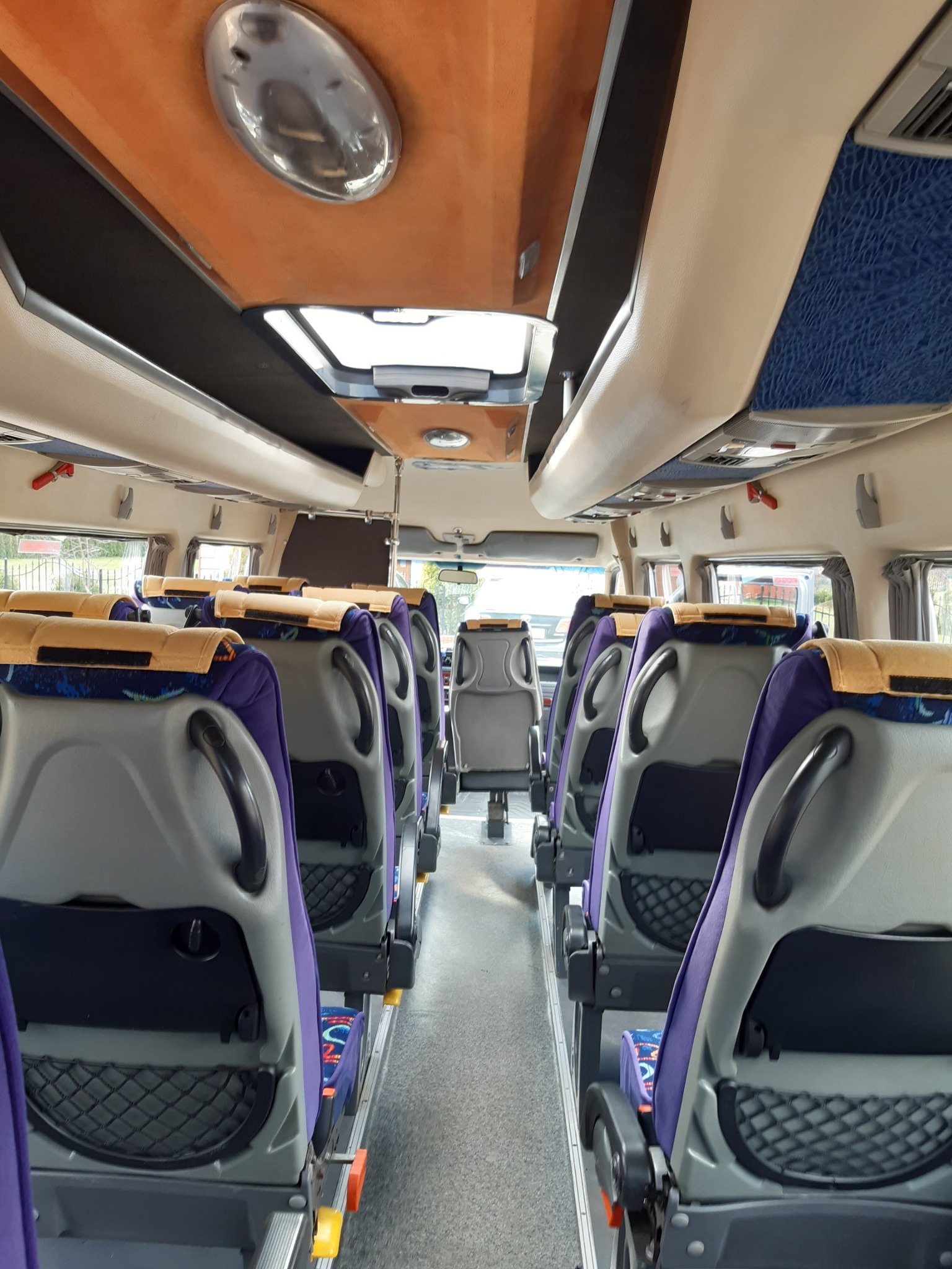 Wnętrze autobusu z rzędami szarych foteli z fioletowymi obiciami i żółtymi zagłówkami, widok z przodu pojazdu, oświetlenie sufitowe i okno dachowe.