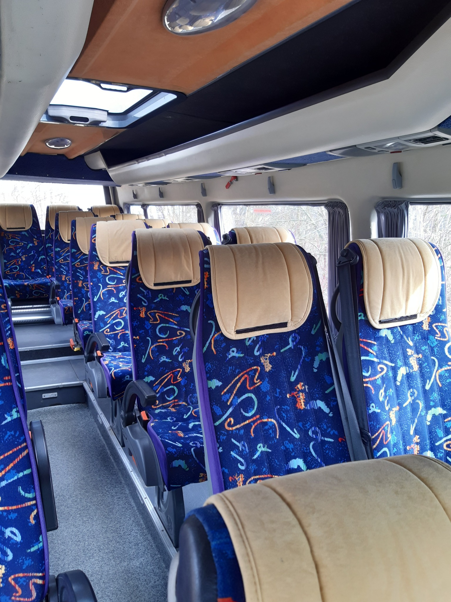 Wnętrze autobusu z rzędem niebieskich foteli z kolorowym wzorem i beżowymi zagłówkami, widok z perspektywy pasażera w kierunku przodu pojazdu, z oknem po prawej stronie.