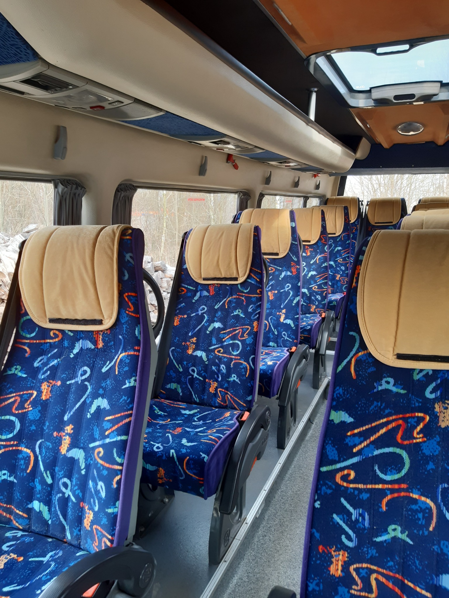Wnętrze autobusu z rzędem foteli obitych niebieską tapicerką ze wzorem, beżowe zagłówki, widok na okno i drzewa w tle.