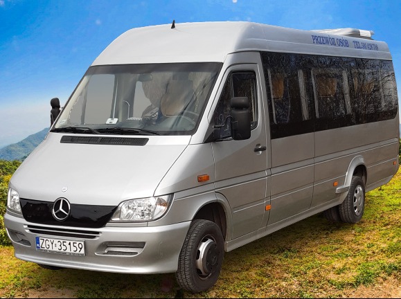 Srebrny bus Mercedes-Benz z napisem 'PRZEWÓZ OSÓB' i numerem telefonu na dachu, zaparkowany na trawiastym terenie z widokiem na góry w tle.