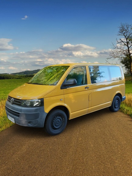 Żółty van Volkswagen Transporter z czarnymi felgami na polnej drodze z widokiem na łąki i lasy.
