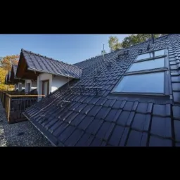 Ciemna dachówka ceramiczna na skośnym dachu z trzema oknami połaciowymi, widok z dołu na budynek z drewnianą balustradą balkonu w tle.