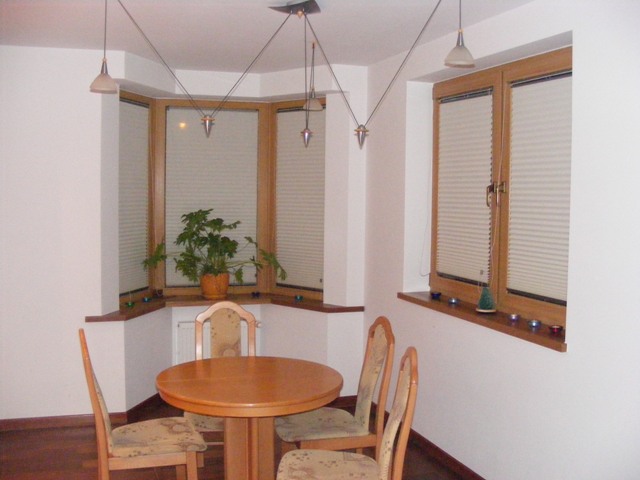 Jadalnia z drewnianymi oknami wyposażonymi w plisowane rolety, okrągły stół i krzesła w neutralnych kolorach, oświetlona wiszącymi lampami.