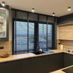 Nowoczesna kuchnia z czarnymi meblami i drewnianym blatem, okno z poziomymi żaluzjami w kolorze grafitowym, widok na zewnątrz z basenem.