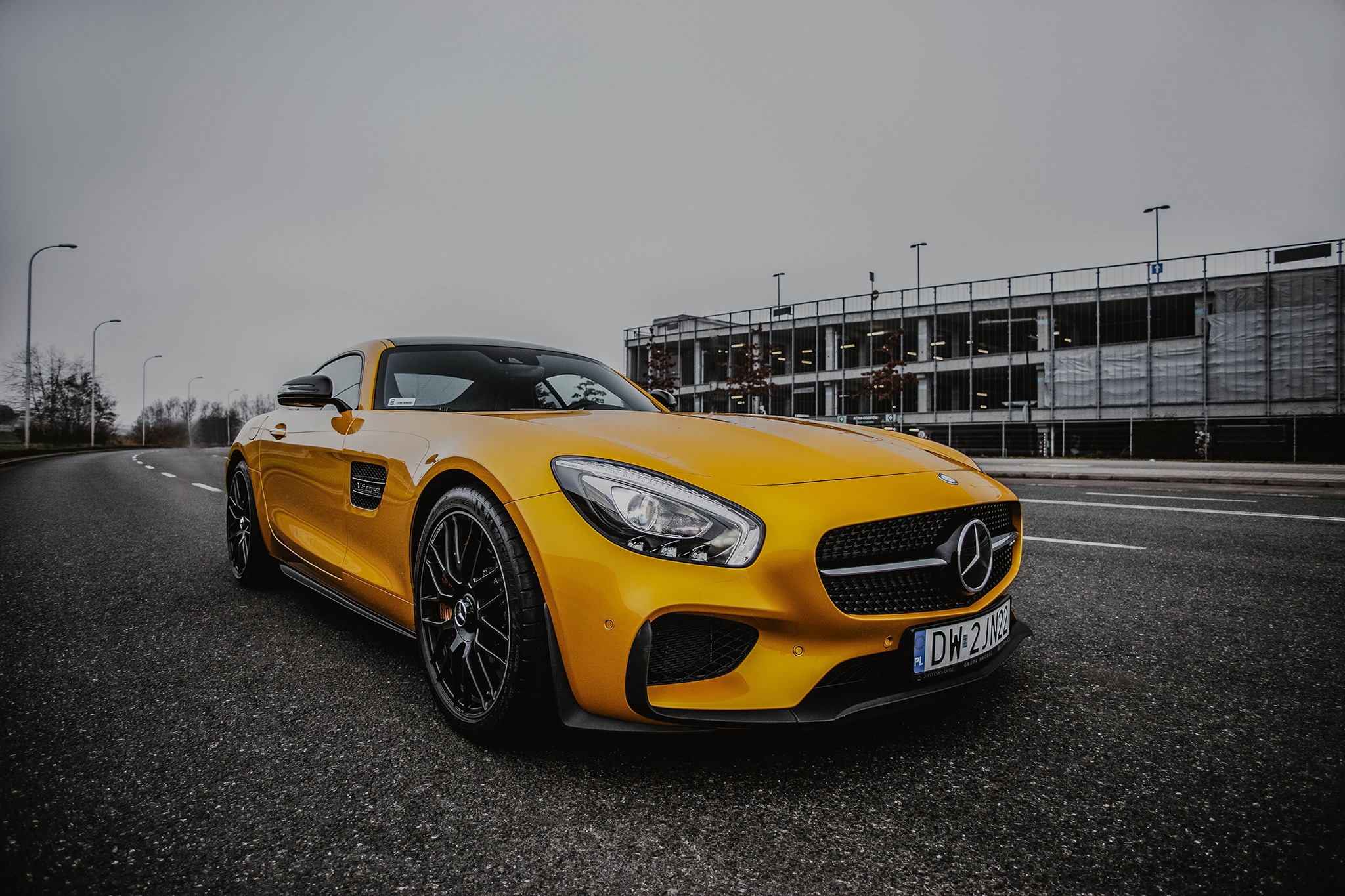 Żółty sportowy Mercedes-AMG GT na asfalcie, z budynkiem parkingu w tle i pochmurnym niebem.