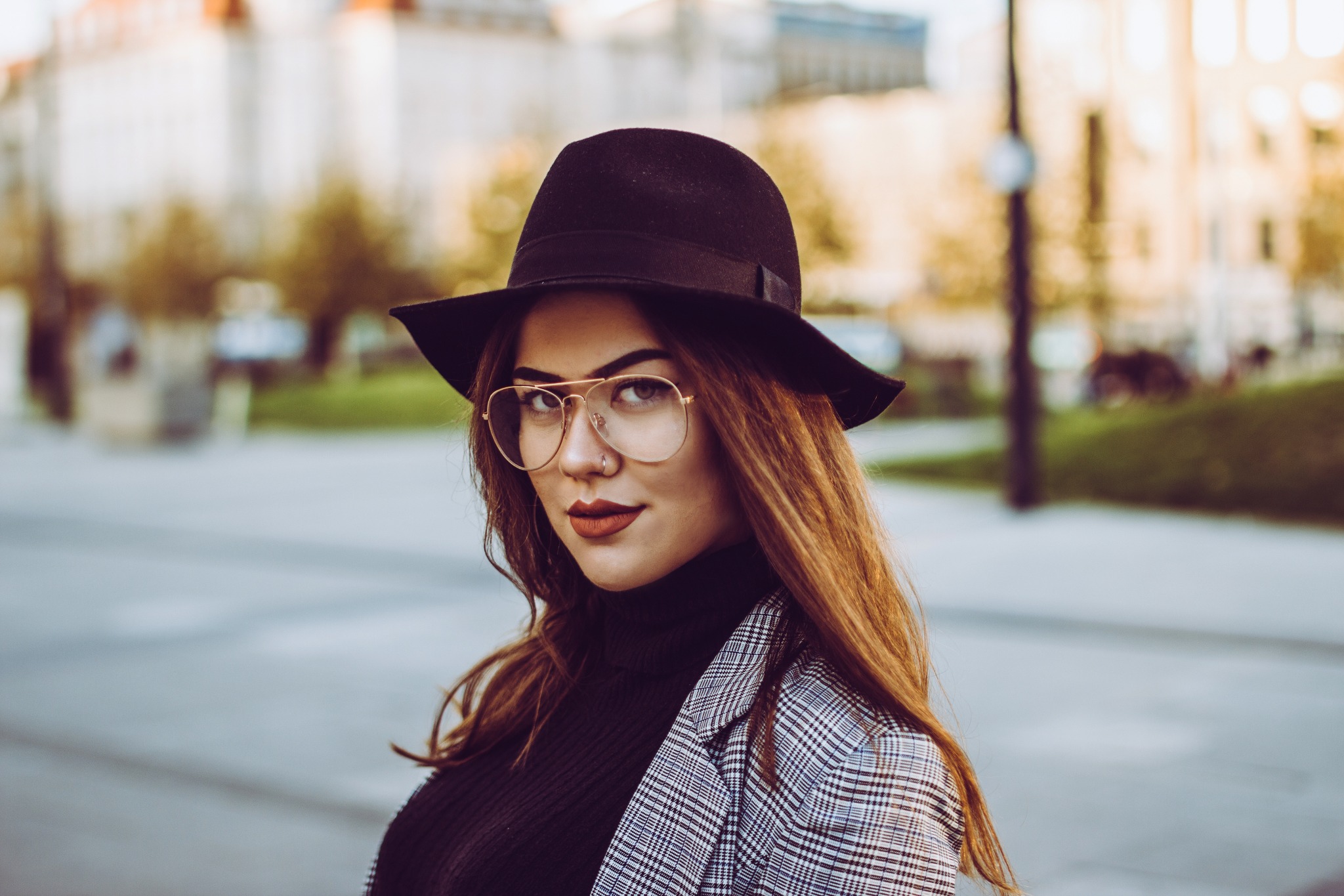 Portret kobiety w czarnym kapeluszu i okularach w złotych oprawkach, ubrana w czarny golf i marynarkę w kratę na tle miejskiego krajobrazu.