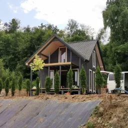 Nowoczesny, ciemny dom z poddaszem użytkowym i drewnianym wykończeniem elewacji, otoczony młodymi tujami i zielenią, z białym busem w tle.