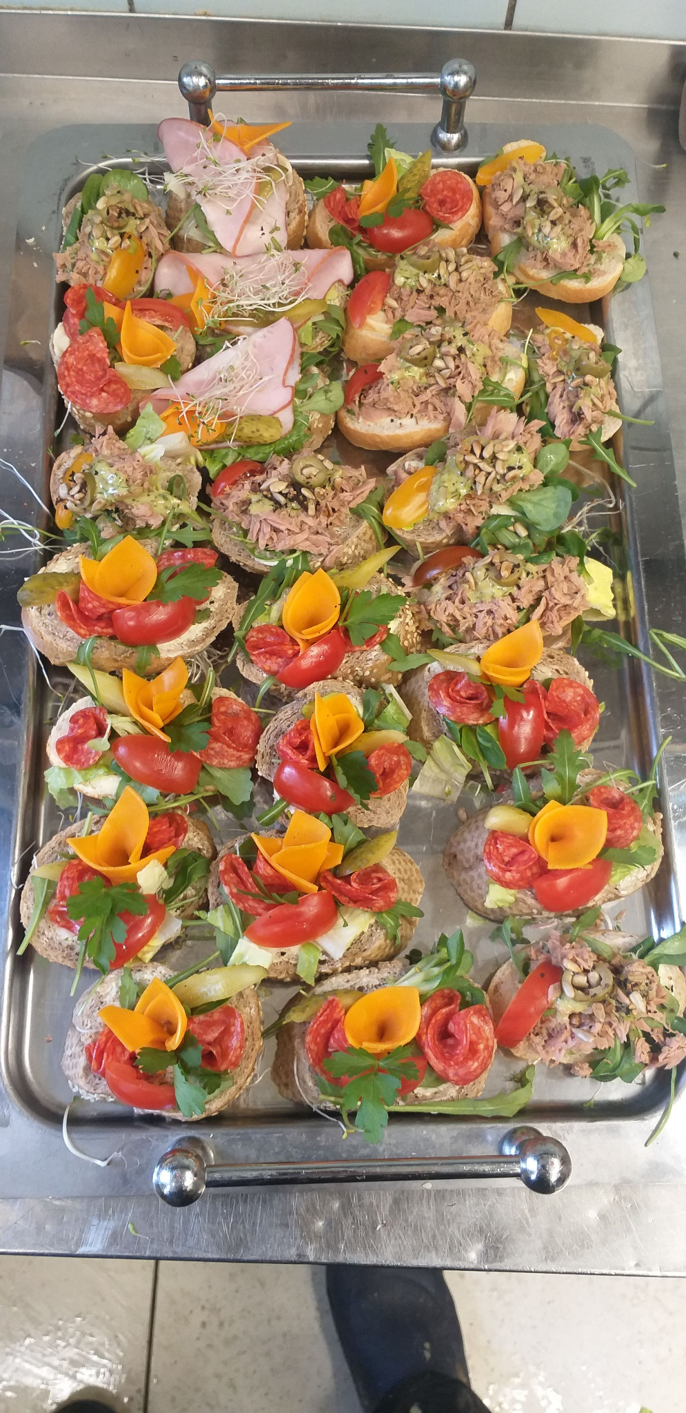 Taca z różnorodnymi kanapkami na ciemnym pieczywie, udekorowanymi wędlinami, warzywami i ziołami, z widocznym fragmentem buta w dolnej części kadru.