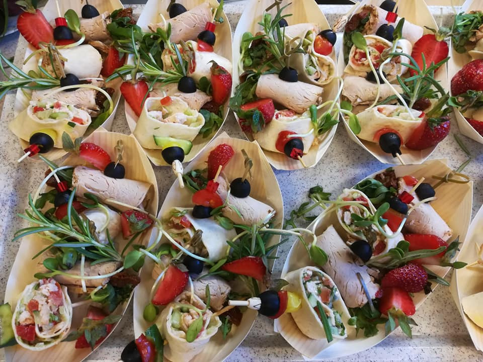 Elegancko podane roladki z tortilli z indykiem, warzywami i truskawkami, udekorowane gałązkami rozmarynu i czarnymi oliwkami na wykałaczkach, w małych drewnianych łódeczkach.