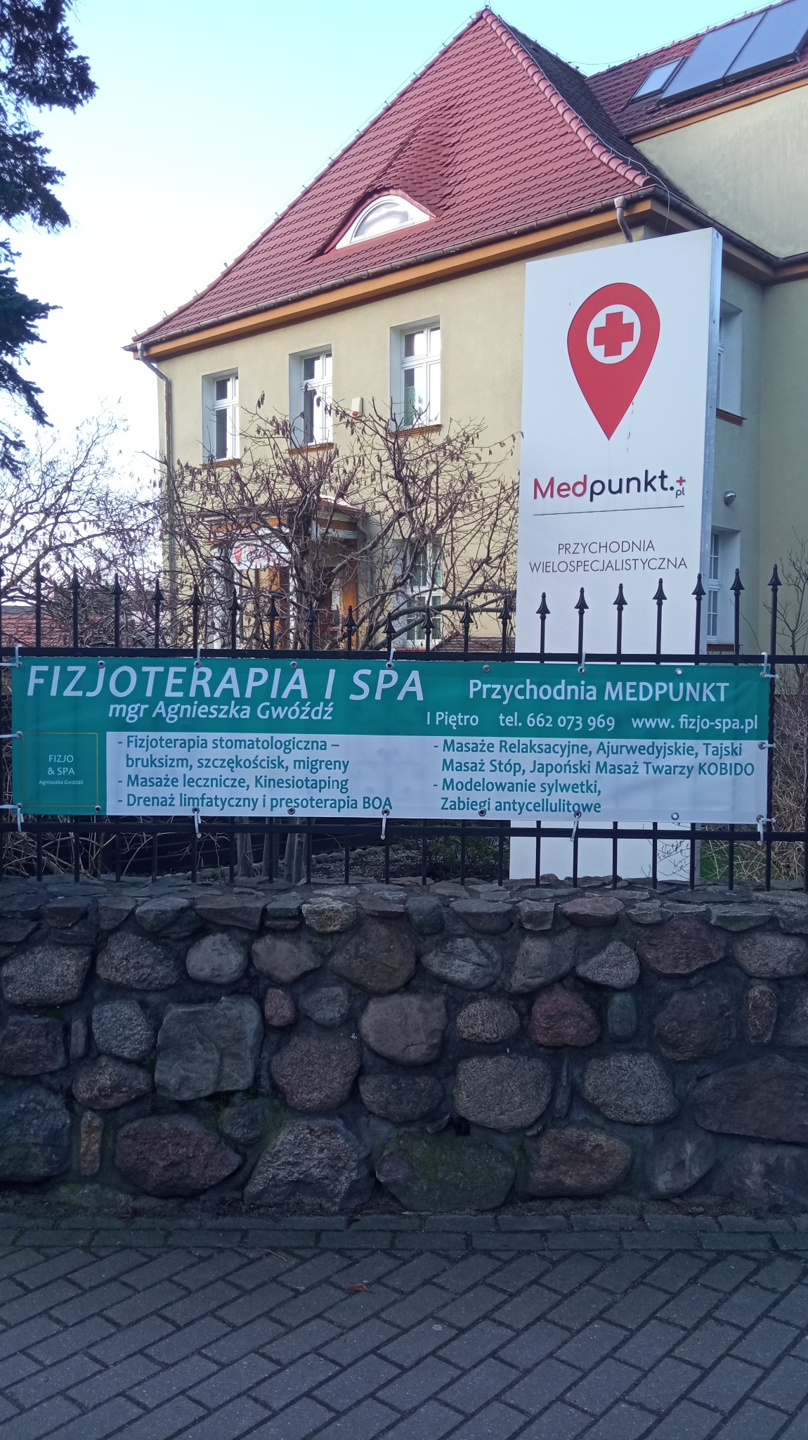 Budynek przychodni Medpunkt z zielonym banerem reklamującym fizjoterapię i spa, z ofertą masaży i zabiegów, widoczny kamienny murek i drzewa bez liści.