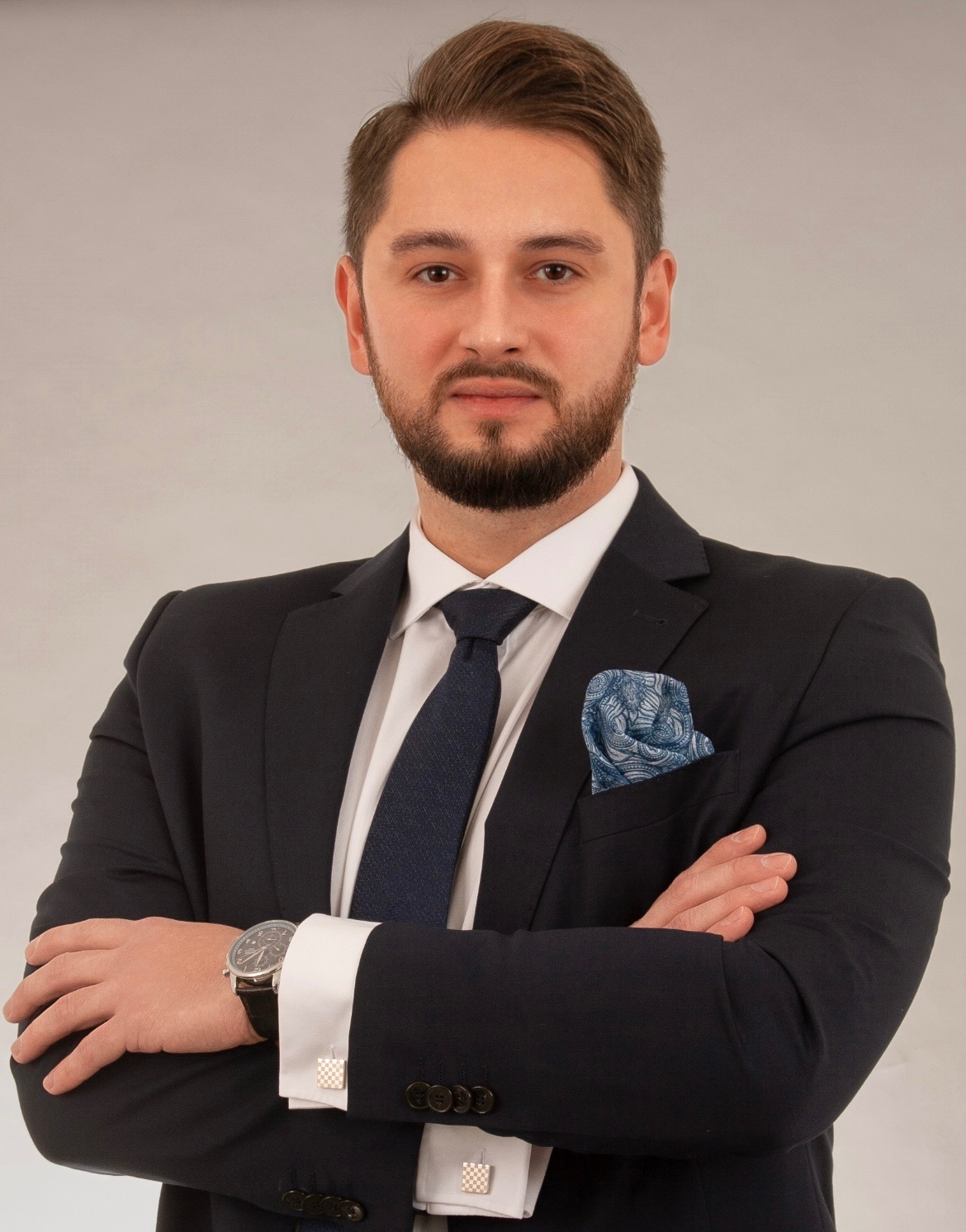 Portret mężczyzny w garniturze z poszetką i zegarkiem, skrzyżowane ręce, profesjonalny wygląd, budzący zaufanie, na jasnym tle.