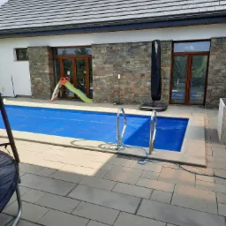 Zewnętrzny basen z niebieską pokrywą, drabinką i zjeżdżalnią dla dzieci, otoczony kamienną elewacją budynku z drewnianymi oknami i szarym dachem.