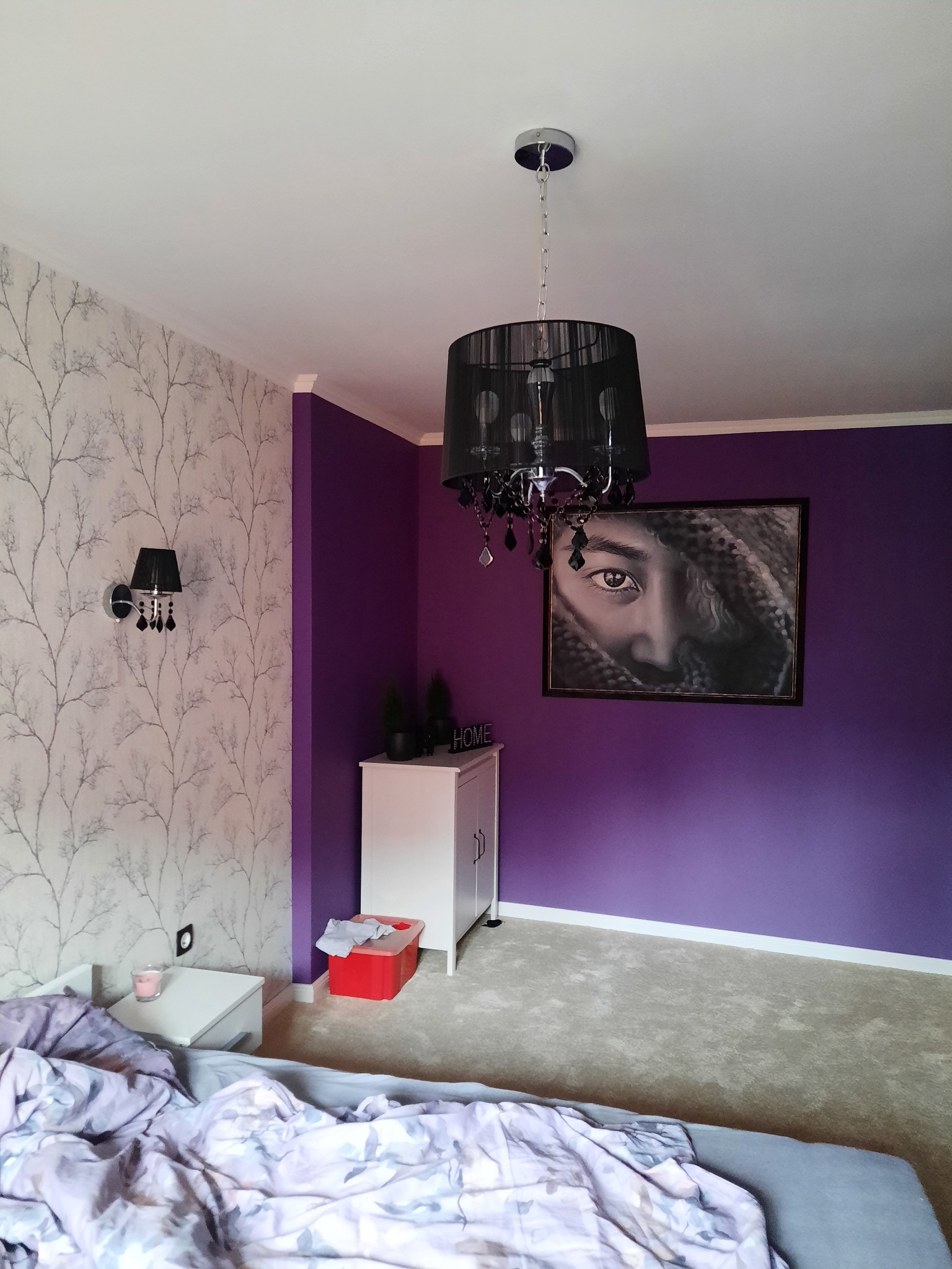 Sypialnia z fioletową ścianą, wzorzystą tapetą, czarnym żyrandolem i obrazem z portretem kobiety.