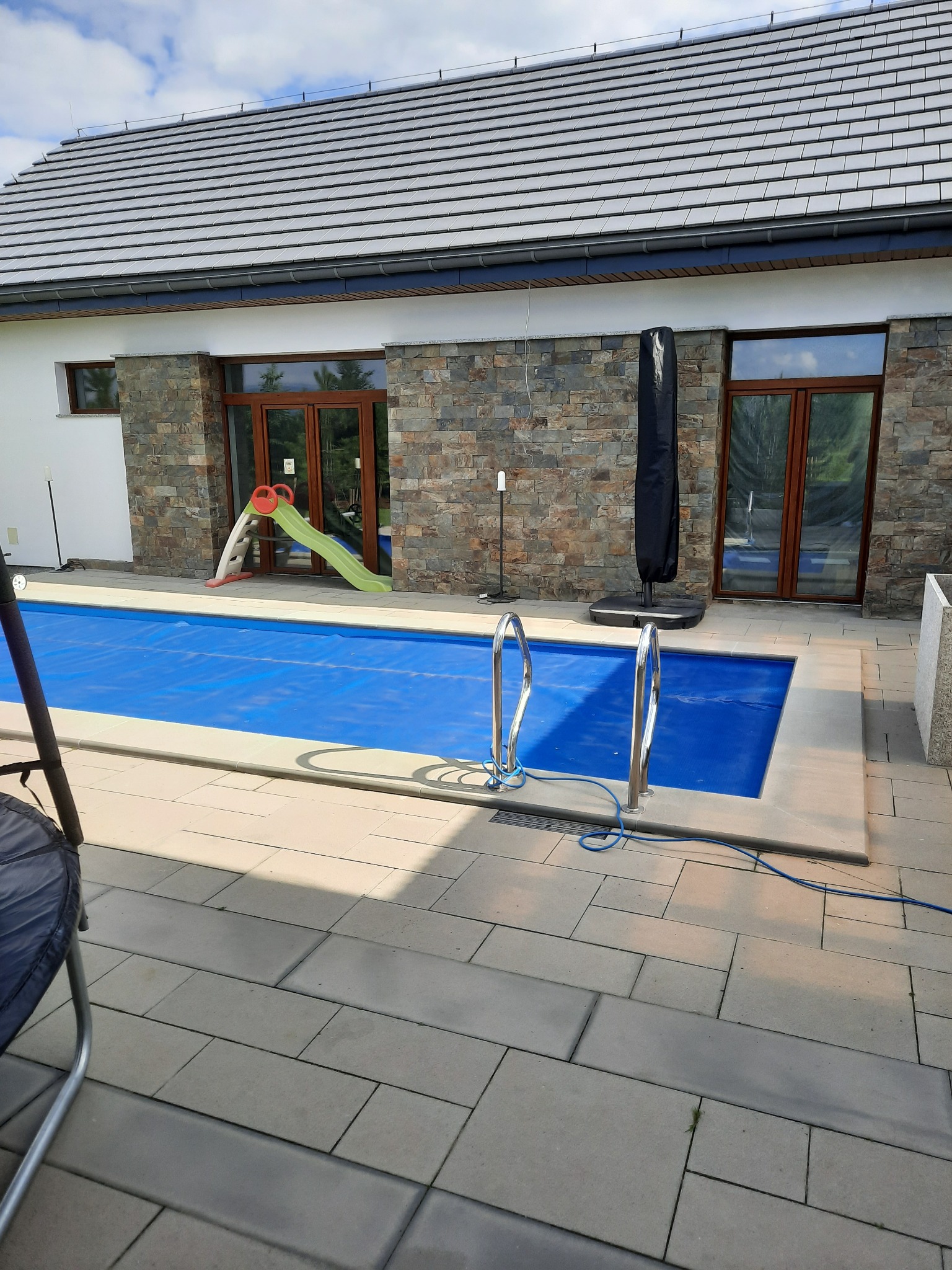 Zewnętrzny basen z niebieską pokrywą, drabinką i zjeżdżalnią dla dzieci, otoczony kamienną elewacją budynku z drewnianymi oknami i szarym dachem.