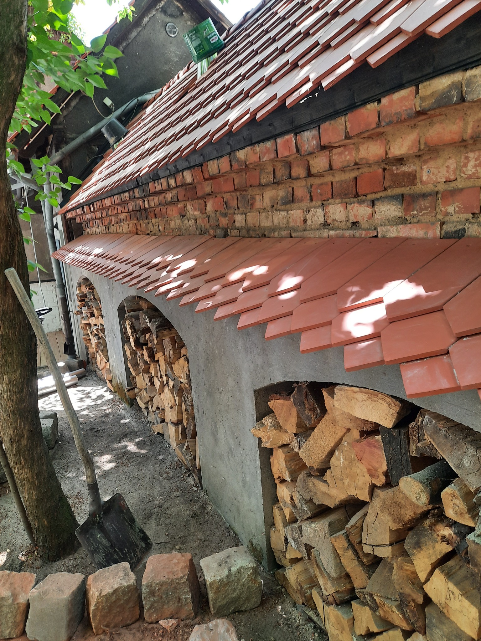 Ceglany budynek z nowym dachem z dachówki ceramicznej, z wnękami wypełnionymi drewnem na opał, obok drzewo i łopata.