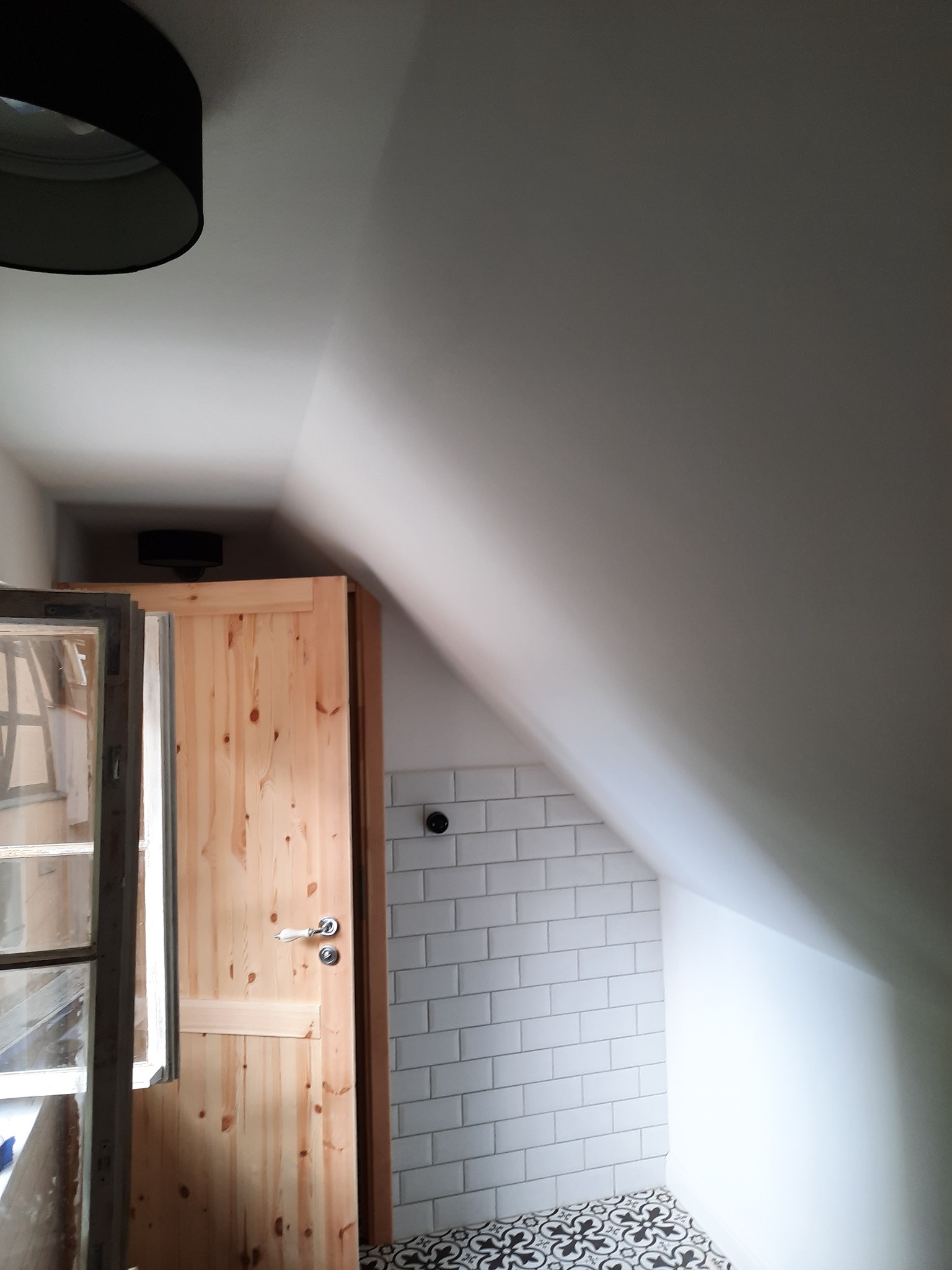 Poddasze po remoncie: białe płytki metra na ścianie, wzorzysta czarno-biała podłoga, drewniane drzwi z oknem, czarny plafon na suficie.