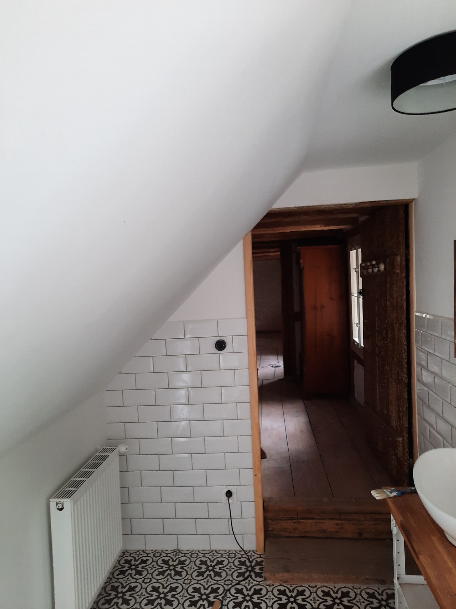 Wykończona łazienka na poddaszu ze skośnym sufitem, białymi płytkami metra na ścianach, wzorzystą podłogą, grzejnikiem i drewnianą framugą drzwi prowadzących do kolejnego pomieszczenia.